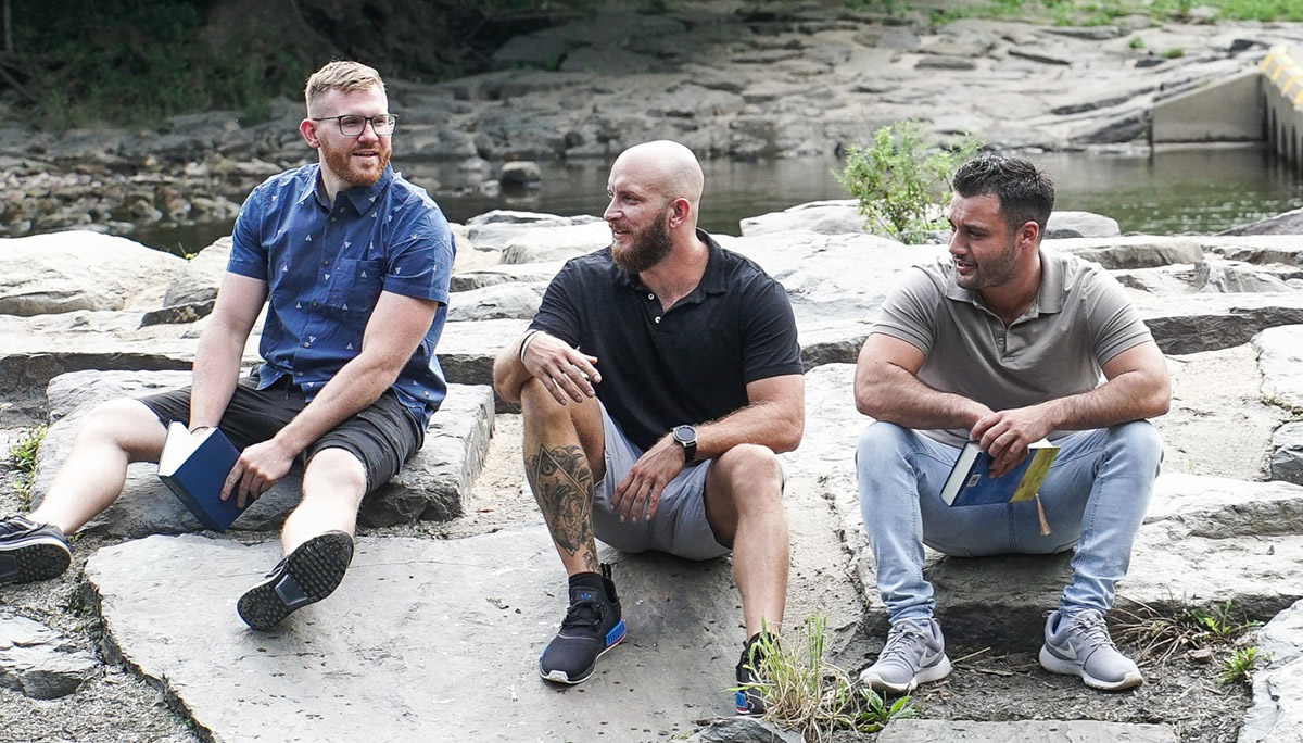 three men sitting on a rock by a river